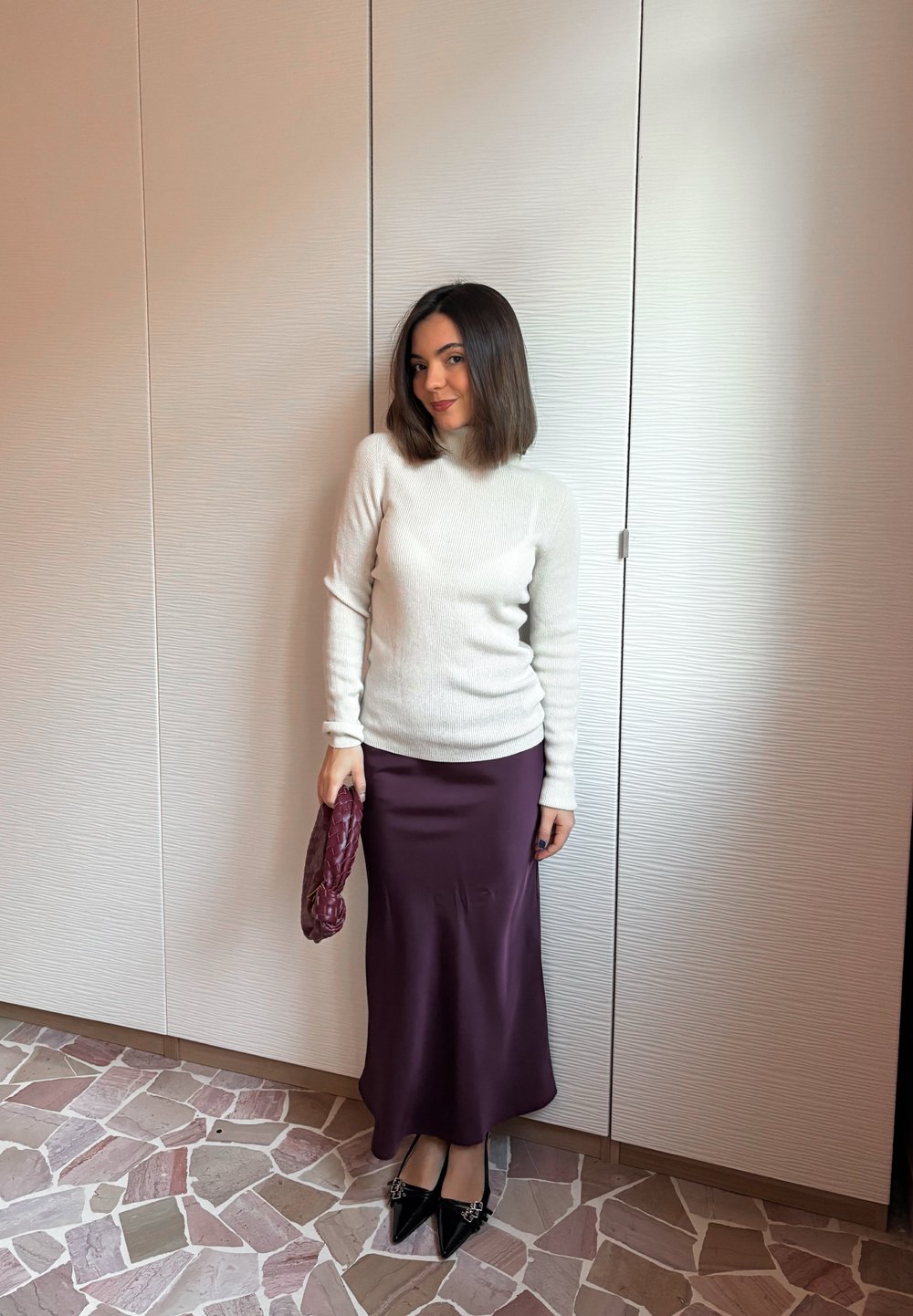 Femme aux cheveux foncés mi-longs portant un pull blanc, une jupe en satin violet, des chaussures noires à bouts pointus, et tenant une pochette violette, debout à l'intérieur.