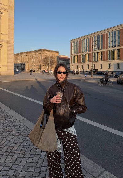 Mujer con gafas de sol oscuras y chaqueta de cuero sostiene una bebida y una gran bolsa beige, de pie en una calle de la ciudad con edificios y ciclistas.
