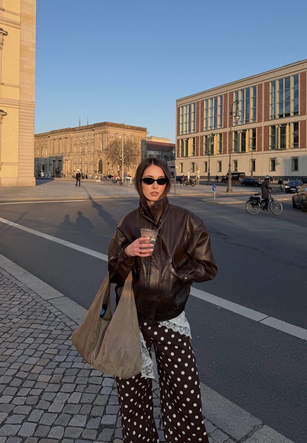 Femme portant des lunettes de soleil foncées et une veste en cuir tient un verre et un grand sac beige, debout dans une rue de la ville avec des bâtiments et des cyclistes.
