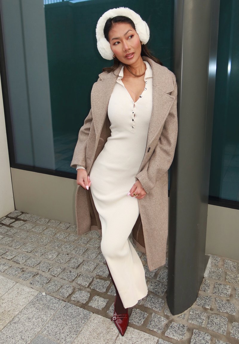 Casaco de lã bege sobre um vestido midi branco canelado com botões, combinado com auriculares brancos e sapatos de salto bordeaux com bico fino, numa superfície de calçada.