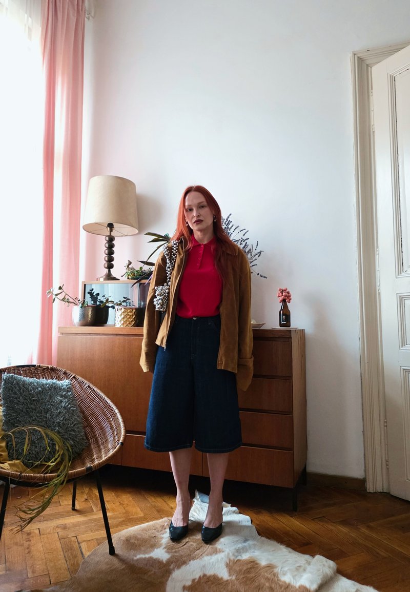 Femme aux cheveux roux debout dans une pièce, portant une chemise rouge, une veste marron, une jupe foncée mi-longue, des talons noirs, à côté d'une armoire en bois et d'une chaise en osier.