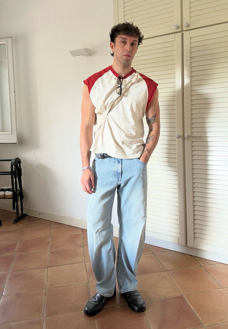 Homme aux cheveux bouclés portant un débardeur blanc et rouge, un jean large bleu clair, des chaussures noires et des lunettes de soleil suspendues à une sangle.