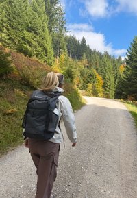 Rucsac negru cu detalii din plasă, purtat peste o jachetă deschisă la culoare și pantaloni maronii, mers pe un drum de pietriș înconjurat de copaci.