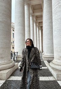 Femme en lunettes de soleil portant un manteau à imprimé léopard et un foulard noir, marchant entre de hautes colonnes en pierre sur un chemin pavé.