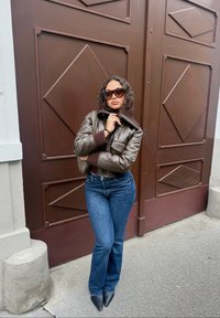 Chaqueta de cuero marrón con cuello alto, suéter de punto oscuro debajo, jeans de campana azul y zapatos negros de punta, contra un fondo de puerta marrón.