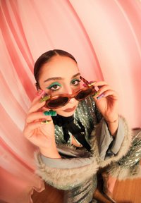 Lunettes de soleil ovales marron avec des verres teintés, tenues par des mains aux ongles colorés et à motifs. Tenue argentée brillante avec une veste bordée de fourrure.