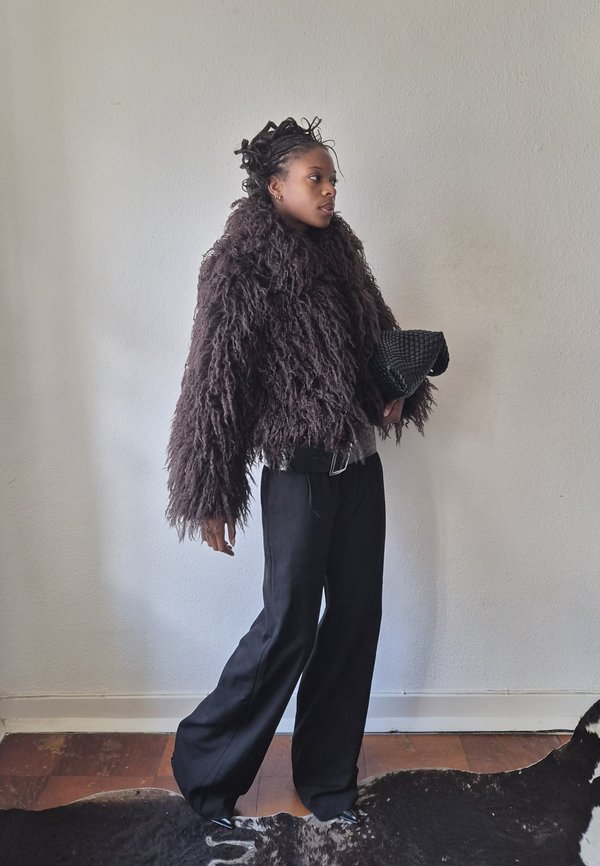Une femme vêtue d’un manteau sombre à l’aspect hirsute et d’un pantalon à jambes larges tient une pochette noire, debout devant un mur uni.