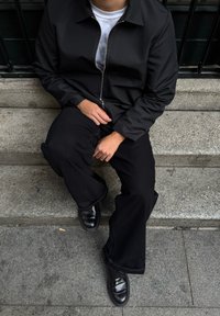 Giacca nera con zip, maglietta bianca sotto e pantaloni neri a gamba larga. Scarpe di pelle nere. Seduto su gradini di pietra. Nessun motivo o accento visibile.