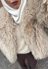 Personne portant un manteau en fausse fourrure beige texturée, un pull en maille crème, une écharpe blanche côtelée à col montant et un pantalon sombre sur un sol pavé.