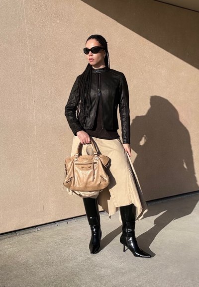 Mujer con chaqueta de cuero negra, falda beige asimétrica, botas negras de tacón alto, gafas de sol, sosteniendo un bolso grande de color beige frente a una pared beige.