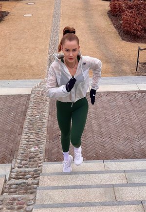 Mujer con chaqueta blanca, mallas verdes y guantes negros corriendo cuesta arriba por unas escaleras de piedra al aire libre con un camino de grava y arbustos secos cerca.