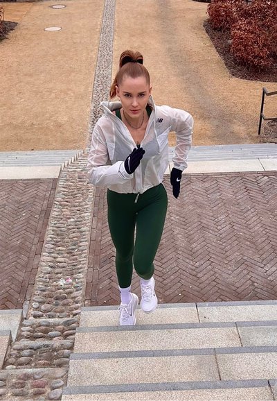 Mujer con chaqueta blanca, mallas verdes y guantes negros corriendo cuesta arriba por unas escaleras de piedra al aire libre con un camino de grava y arbustos secos cerca.