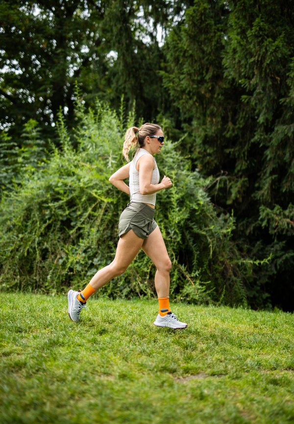 Mujer corriendo sobre hierba en un bosque. Lleva una camiseta sin mangas gris claro, pantalones cortos verde oliva, calcetas naranjas y zapatillas de correr blancas.