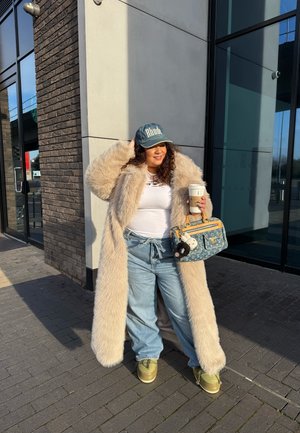 Abrigo de piel sintética marrón claro, blusa blanca, jeans de pierna ancha azul claro, zapatos verde oliva, gorra de mezclilla y bolso con estampado. Taza de café en mano.