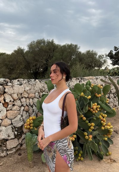 Top blanco sin mangas, falda envolvente con estampado floral y de animal, bolso marrón, pendientes de oro, frente a una pared de piedra y un cactus.