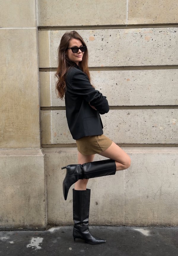 Mujer con gafas de sol, blazer negro, falda beige y botas negras altas posa contra una pared de piedra, doblando una pierna detrás de ella.