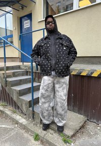 Muž v černé vzorované bundě, šedých kalhotách s tie-dye, černé kapuci a slunečních brýlích stojí s rukama v kapsách vedle betonových schodů.