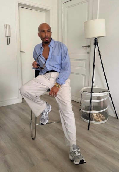 Camisa azul claro abotonada, pantalones beige y zapatillas grises. Sentado en una silla metálica. De fondo, una lámpara de pie y una cesta de almacenamiento.