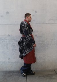 Crni i bijeli karirani oversized kaput s crvenim kožnim detaljima i remenom, nošen preko crnih hlača i masivnih crnih cipela sa srebrnim naglascima.