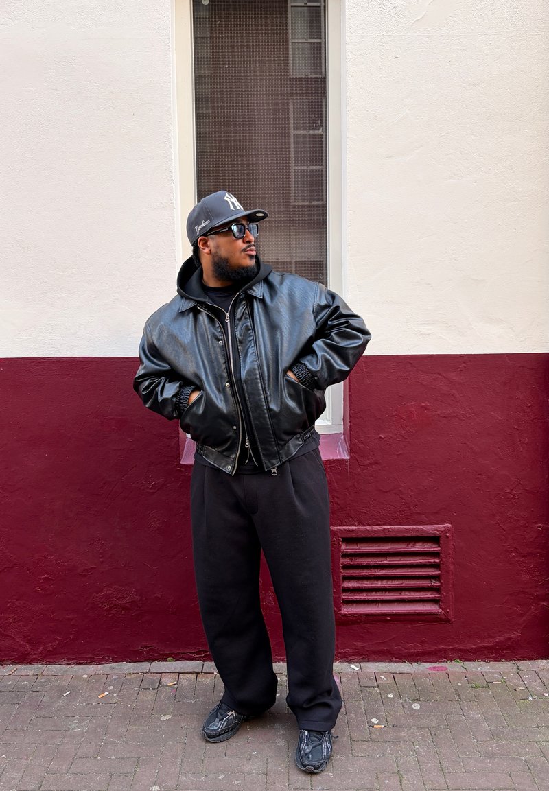 Homme portant une veste en cuir noire, un pantalon noir, des baskets, des lunettes de soleil et une casquette de baseball grise, debout devant un mur blanc et bordeau.
