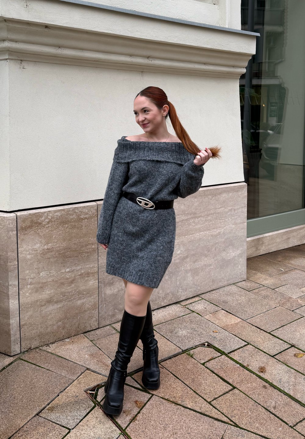 Robe grise tricotée à épaules dénudées, de coupe ajustée, accompagnée d'une large ceinture noire avec une boucle argentée, et de bottes noires à talons montant jusqu'aux genoux.