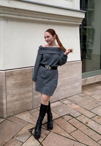 Robe grise tricotée à épaules dénudées, de coupe ajustée, accompagnée d'une large ceinture noire avec une boucle argentée, et de bottes noires à talons montant jusqu'aux genoux.
