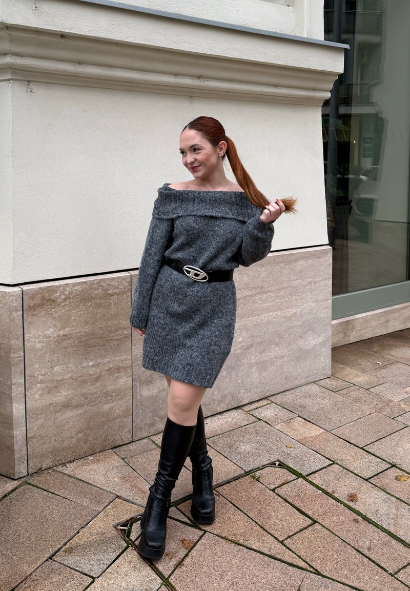 Robe grise tricotée à épaules dénudées et coupe ajustée, associée à une large ceinture noire avec une boucle argentée, et des bottes noires montantes à talons jusqu'au genou.