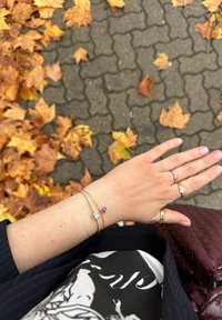 Bracciali d'oro impilati su una mano, con una pietra chiara rettangolare e una pietra rosa a forma di cuore. Foglie e un sentiero testurizzato visibili sullo sfondo.