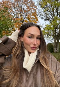 Brun skinnfrakk med høy krage, kombinert med et kremfarget, strikket skjerf. Langt brunt hår og synlig utendørs løvverk i høstfarger.