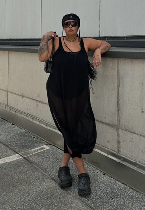 Vestido negro transparente sin mangas con aberturas laterales, combinado con Crocs negros. Los accesorios incluyen un bolso pequeño y gafas de sol. Fondo de hormigón.