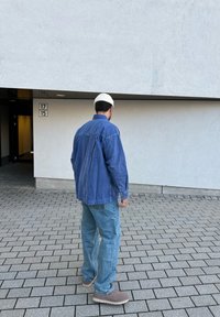 Camisa de denim azul com mangas longas, em corte descontraído, combinada com jeans azul claro e sapatos cinzentos, usada com um chapéu de malha branco.