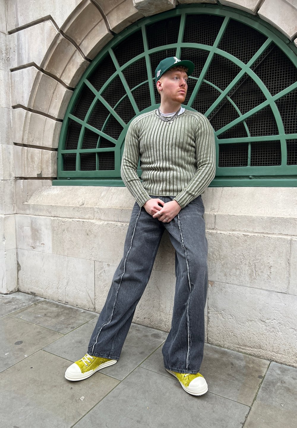 Green ribbed sweater, gray wide-leg jeans with white stitching, and yellow sneakers. Background features an arched green window.