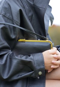 Geantă de mână din piele neagră cu închidere aurie, ținută sub o geacă de piele neagră. Designul este elegant, cu o textură netedă.