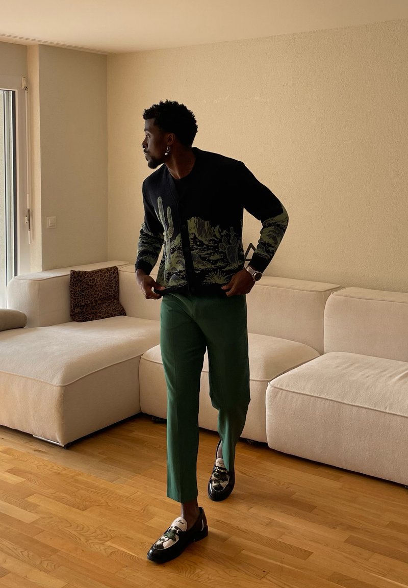 Man adjusting black cactus-patterned cardigan while standing on wooden floor near beige sectional sofa with leopard-print pillow.