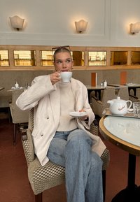 Fehér kerámia csésze jobb kézben tartva, mellé egy kis fehér csészealj. Világos bézs színű, túlméretezett blézer, krémszínű kötött pulóver és kék farmer, asztalnál ülve.