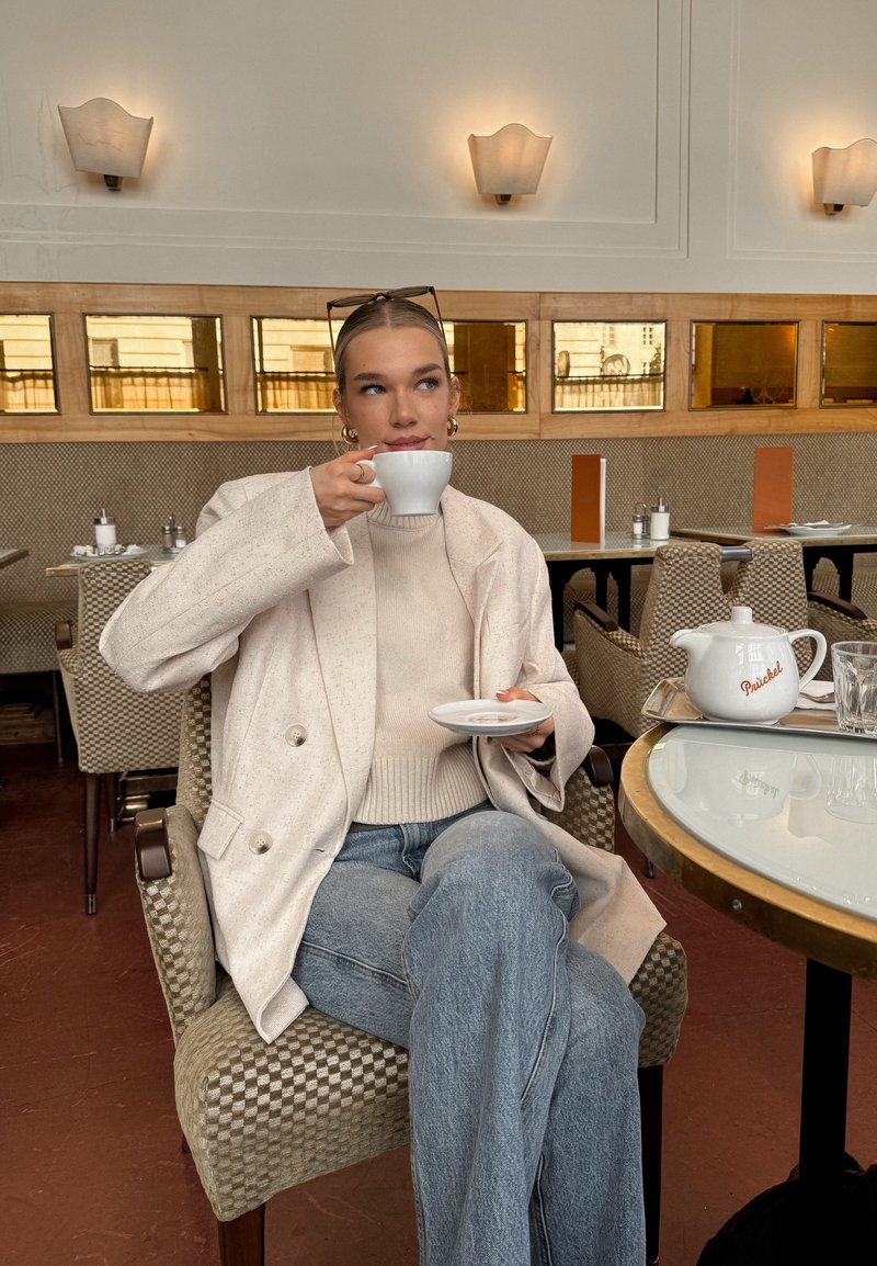 Weiße Keramiktasse in der rechten Hand gehalten, kombiniert mit einer kleinen weißen Untertasse. Hellbeiger übergroßer Blazer, cremefarbener Strickpullover und blaue Jeans, sitzend an einem Tisch.