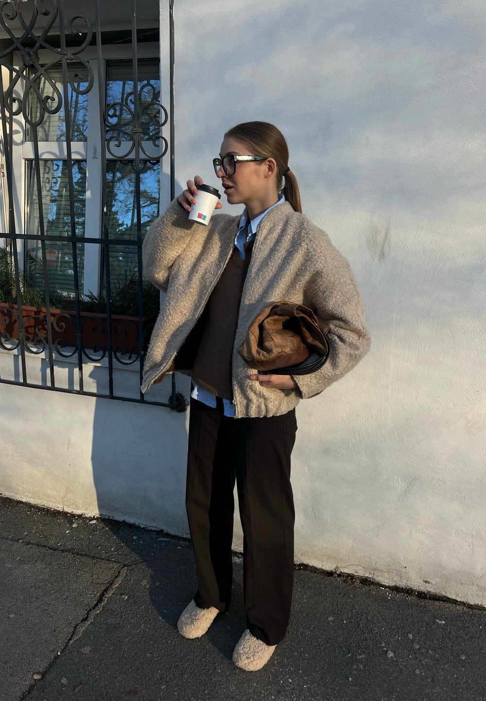 Μπεζ υφασμάτινη μπουφάν, καφέ πουλόβερ, μπλε πουκάμισο, μαύρα ριγέ παντελόνια και μπεζ φλιτζάκια παντόφλες. Κρατάει ένα φλιτζάνι καφέ.