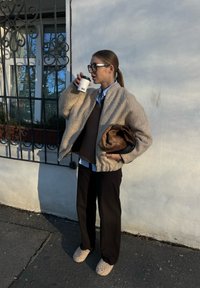 Μπεζ υφασμάτινη μπουφάν, καφέ πουλόβερ, μπλε πουκάμισο, μαύρα ριγέ παντελόνια και μπεζ φλιτζάκια παντόφλες. Κρατάει ένα φλιτζάνι καφέ.