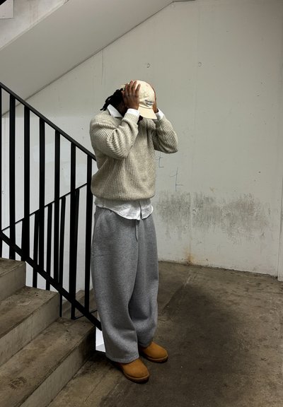 Suéter de punto beige sobre una camisa blanca de botones, pantalones de chándal grises y zuecos marrones. De pie junto a una barandilla negra en un espacio interior minimalista.