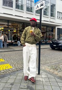 Homme portant une casquette rouge, une veste olive, un pantalon blanc et des chaussures marron, tenant une tasse de café, debout sur un trottoir pavé près d'un panneau de signalisation.