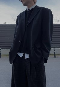 Personne portant un blazer noir oversize, un pantalon large à carreaux sombre, une chemise blanche, un pull noir et une cravate à l'extérieur, les mains dans les poches.