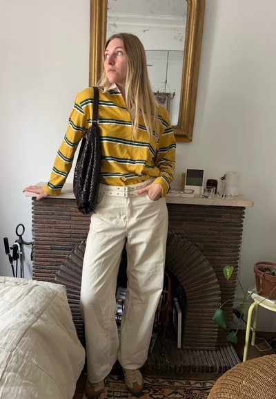 Camisa de rayas amarilla y verde de manga larga, pantalones anchos beige, bolso negro de mimbre, de pie frente a una chimenea de piedra texturizada.