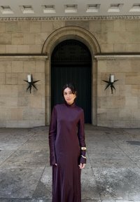 Vestito a maniche lunghe con collo alto in raso bordeaux scuro dalla texture liscia. Presenta un accento dorato unico sulla manica sinistra. Sfondo urbano.