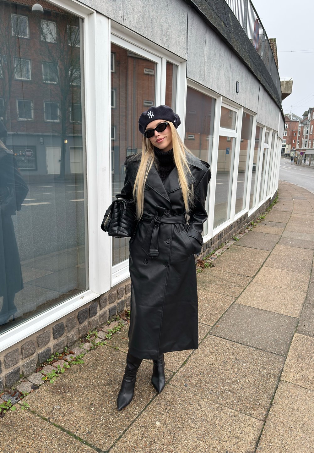 Femme portant un manteau en cuir noir, un béret noir, des lunettes de soleil et des bottes, debout sur un trottoir à côté d'un bâtiment avec de grandes fenêtres.