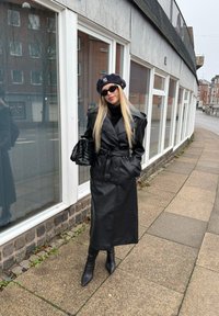 Femme portant un manteau en cuir noir, un béret noir, des lunettes de soleil et des bottes, debout sur un trottoir à côté d'un bâtiment avec de grandes fenêtres.