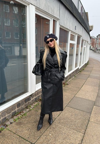Mujer con abrigo de cuero negro, boina negra, gafas de sol y botas, de pie en una acera junto a un edificio con grandes ventanas.