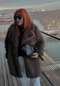 Manteau en fourrure marron, col roulé noir, sac à main noir avec ferrures en or rose, pantalon blanc et lunettes de soleil noires surdimensionnées. Vue de la ville en arrière-plan.