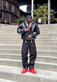 Czarna, skórzana, oversize'owa kurtka z elastycznymi mankietami, biała koszula z kołnierzykiem, czerwona krawat, czarne luźne spodnie oraz jasnoczerwone buty. Przy spodniach breloczki.