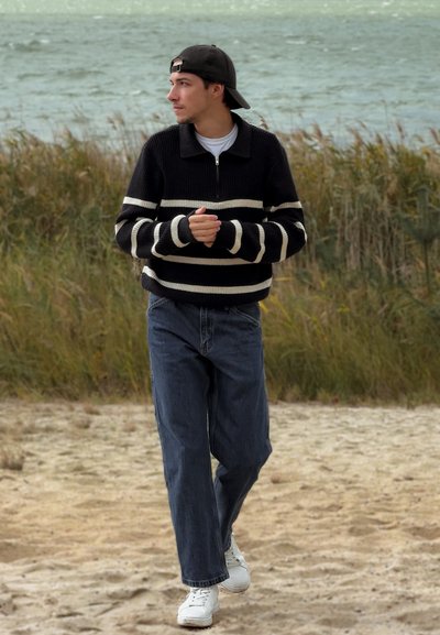 Jersey a rayas blanco y negro con cuello y cremallera, combinado con unos vaqueros oscuros holgados y zapatillas blancas, utilizado al aire libre con un fondo natural.