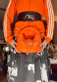 Kirkas oranssi nahkalaukku, jossa on pyöreä muoto ja etuosassa teksturoitu käärmeennahkakuosi, jota pitää kädessään henkilö oranssissa takissa.